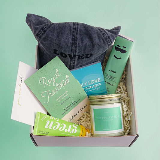 Gift box with various items including a candle, cards, and a hat on a light green background